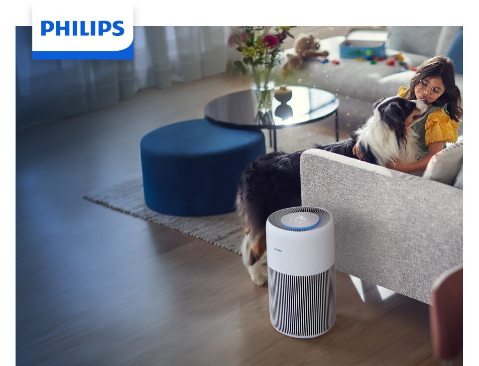 Na obrazie widać oczyszczacz powietrza marki PHILIPS ustawiony obok kanapy w salonie. W tle znajduje się kobieta siedząca z psem na kanapie oraz stolik z kwiatami.