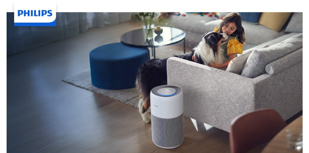 Na obrazie widać oczyszczacz powietrza marki PHILIPS stojący obok kanapy, na której siedzi osoba z psem. W tle znajduje się stolik z kwiatami i pufą, a na górze widoczne jest logo PHILIPS.
