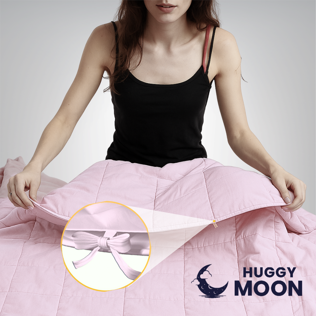 Osoba trzyma różową kołdrę z widocznym zbliżeniem na wiązanie. W prawym dolnym rogu znajduje się logo z napisem \'HUGGY MOON\'.