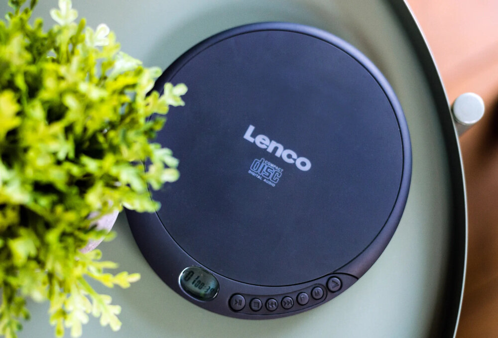 Na okrągłym stoliku znajduje się czarny Discman z widocznym logo \'Lenco\' oraz napisem \'disc digital audio\'. Obok urządzenia widoczna jest zielona roślina.