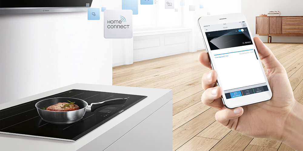Na obrazie widać płytę indukcyjną z garnkiem, w którym znajduje się jedzenie. Obok osoba trzyma smartfon z aplikacją, a w tle widoczne jest logo \'Home Connect\'.