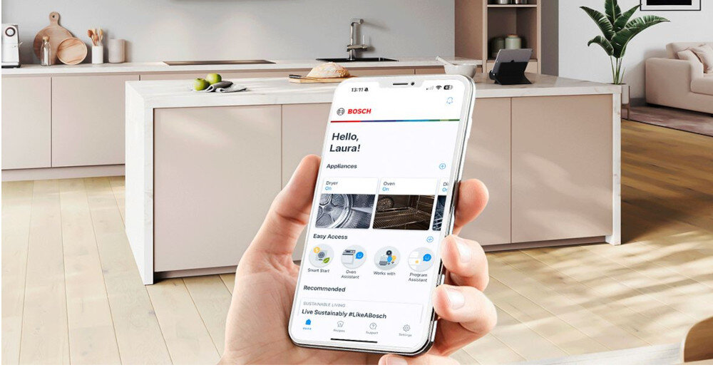 Osoba trzyma smartfon z aplikacją, na której widoczny jest tekst \'Hello, Laura!\' oraz logo BOSCH. W tle znajduje się nowoczesna kuchnia z wyspą i rośliną doniczkową.