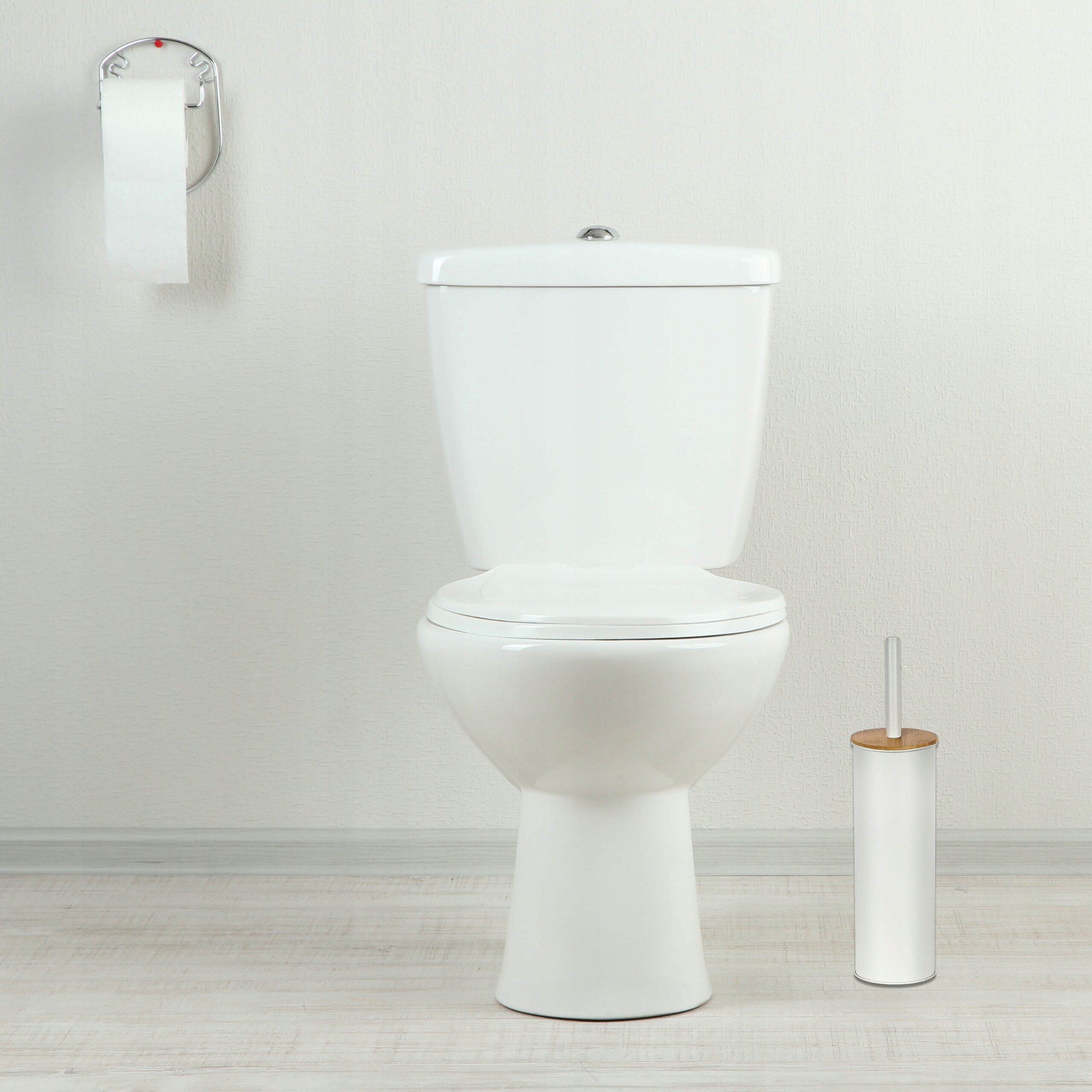 Na obrazie znajduje się biały sedes oraz wolnostojąca szczotka do WC w białej, matowej tubie z drewnianym elementem. Obok sedesu wisi uchwyt z rolką papieru toaletowego.