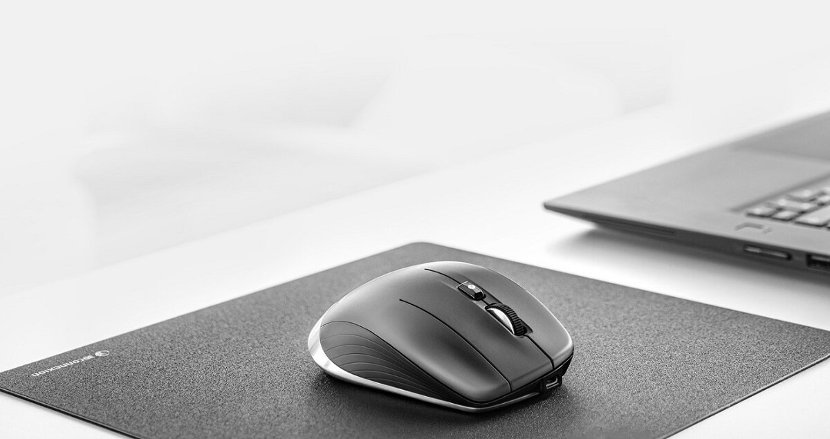 Na obrazie widoczna jest czarna mysz komputerowa umieszczona na podkładce obok laptopa. W lewym dolnym rogu podkładki znajduje się logo \'3Dconnexion\'.