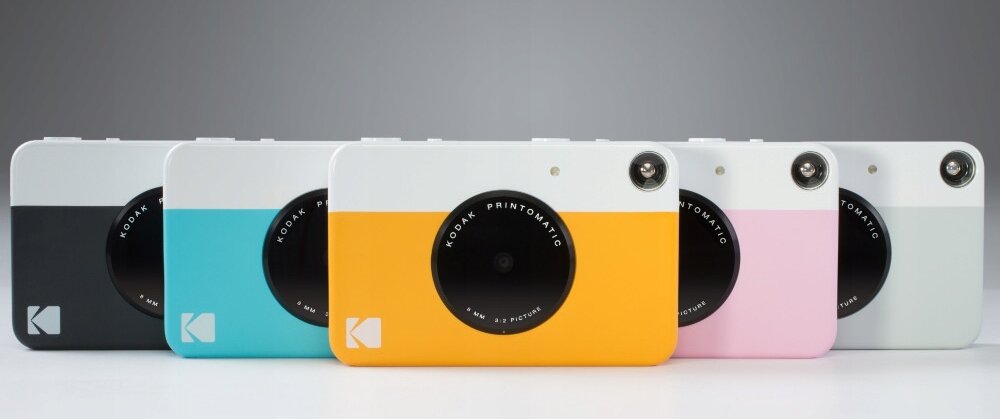 Na obrazie widoczne są aparaty natychmiastowe w różnych kolorach: czarnym, niebieskim, żółtym, różowym i szarym. Każdy aparat ma logo \'KODAK\' oraz napis \'KODAK PRINTOMATIC\' wokół obiektywu.