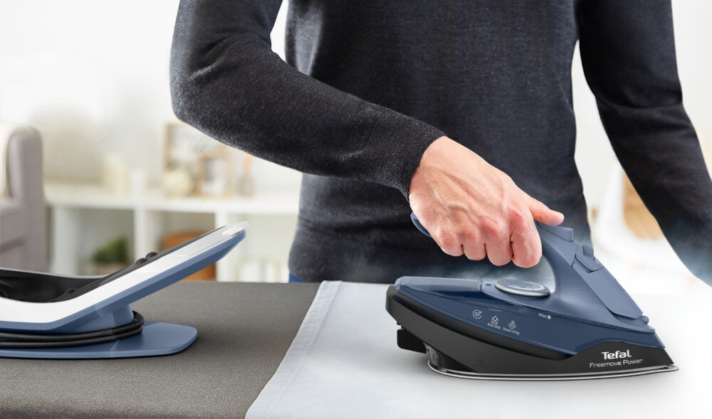 Osoba prasuje tkaninę za pomocą żelazka w kolorze niebieskim z widocznym logo \'Tefal Freemove Power\'. Obok znajduje się stacja dokująca na szarej powierzchni.