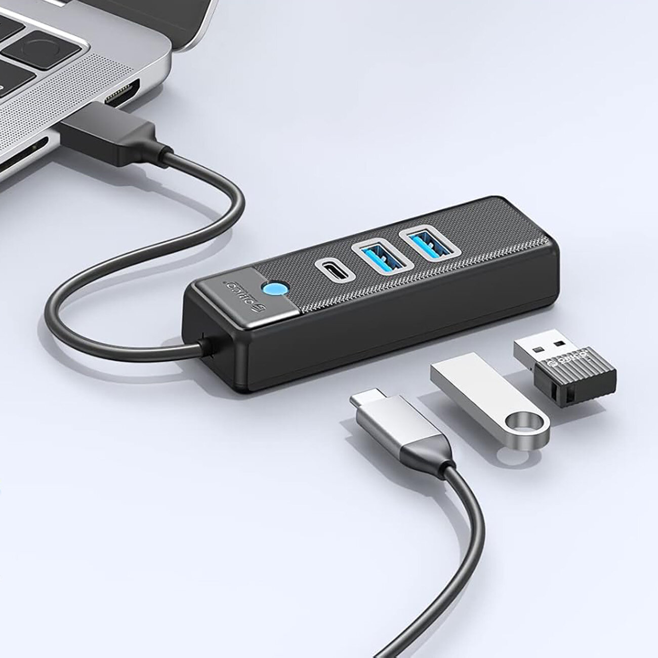 Na obrazie widoczny jest czarny hub USB z dwoma portami USB-A 3.0 i jednym portem USB-C 3.0, podłączony do laptopa. Obok huba leżą trzy różne wtyczki USB, a na urządzeniu widnieje logo \'Orico\'.