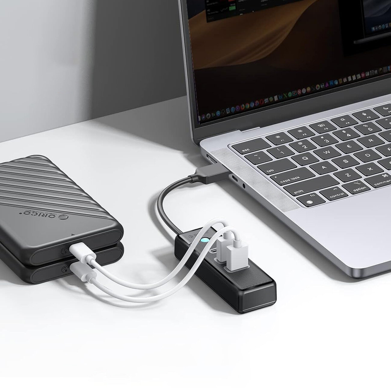 Na obrazie widać laptopa podłączonego do czarnego huba USB, do którego podłączone są dwa białe kable. Obok znajduje się czarne urządzenie z logo \'Orico\'.