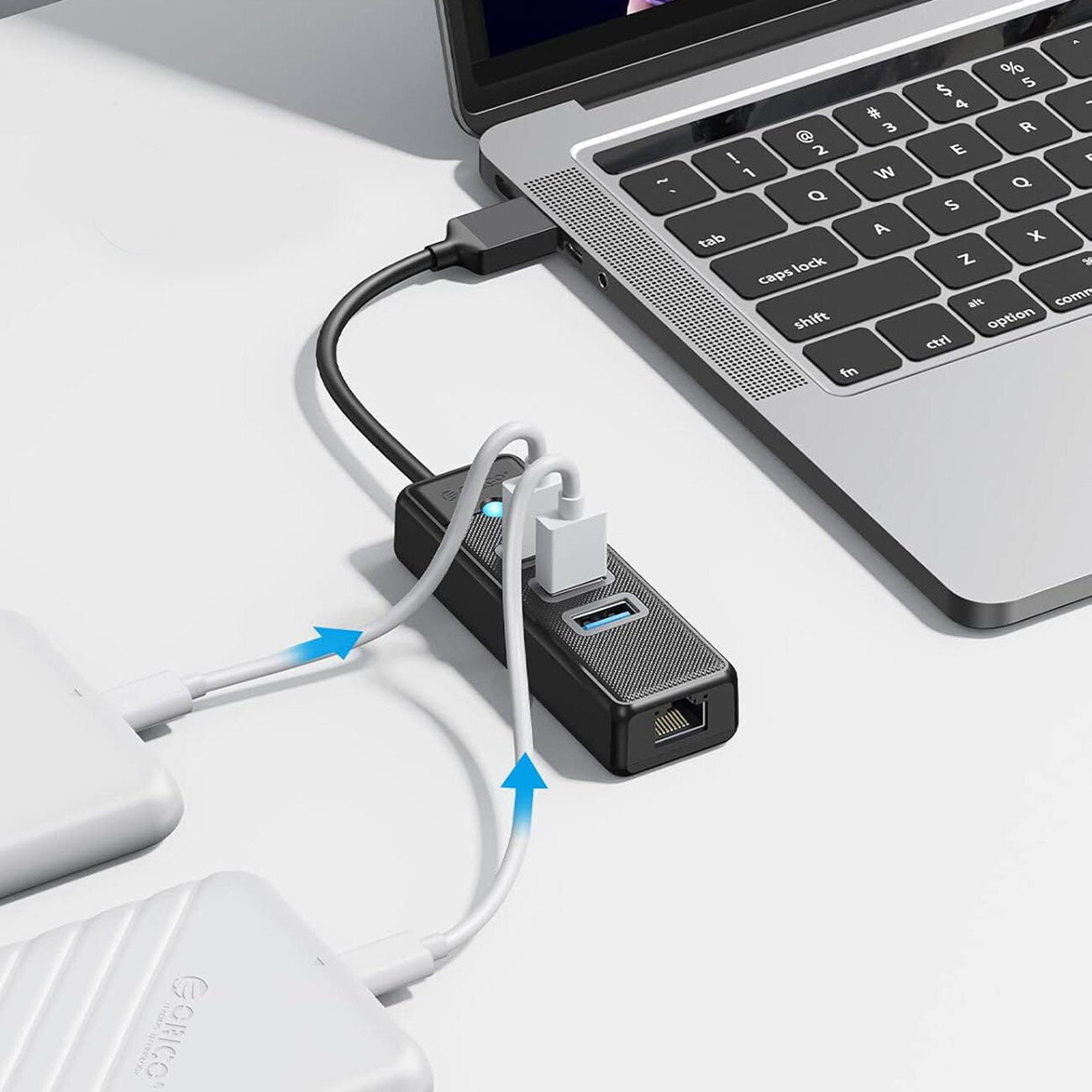 Na obrazie widoczny jest czarny hub USB podłączony do laptopa, z kilkoma kablami wpiętymi w porty USB i Ethernet. Na hubie znajduje się logo \'Orico\'.