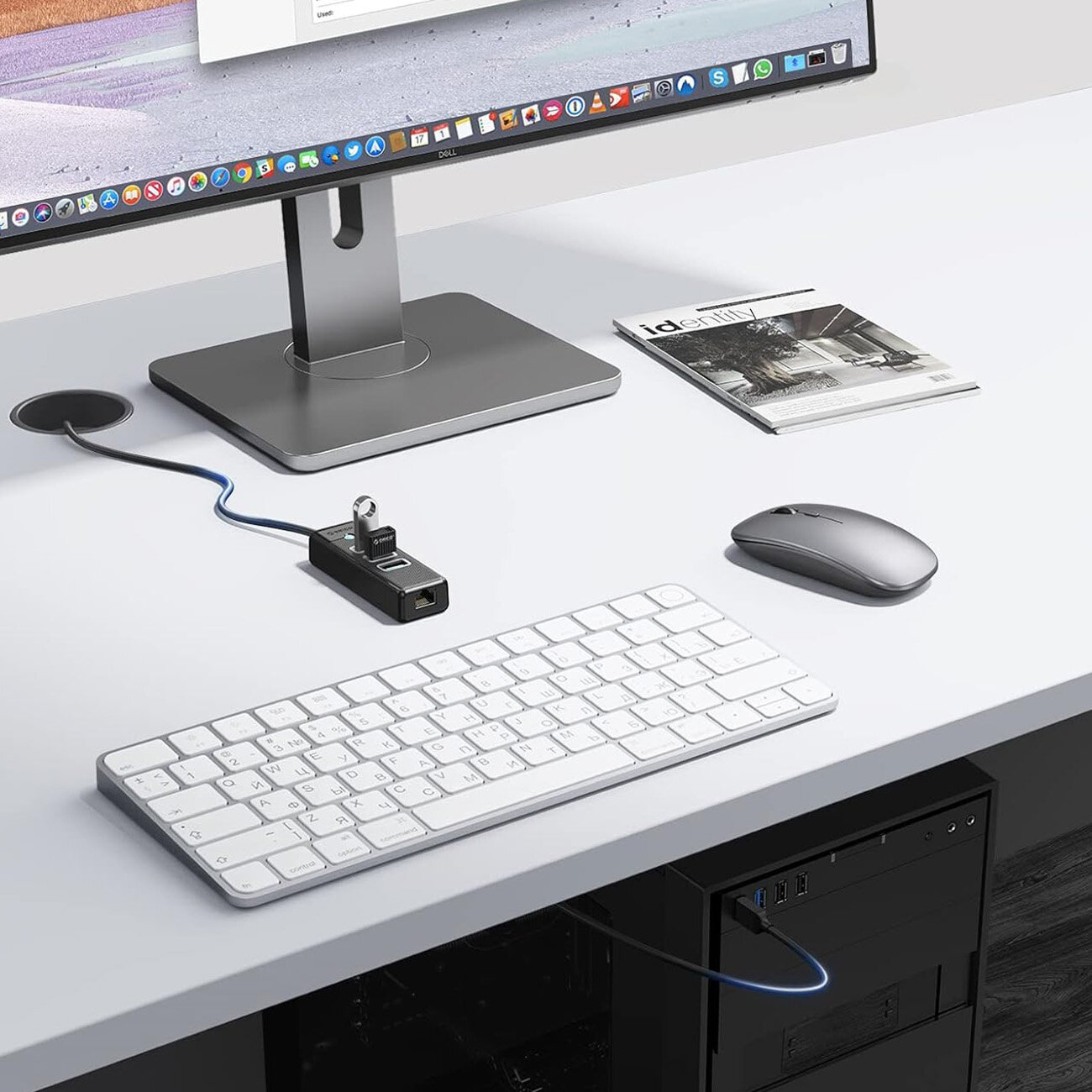 Na biurku znajduje się monitor, klawiatura, mysz oraz czarny hub USB z trzema portami USB-A i jednym portem Ethernet. Obok monitora leży magazyn z widocznym napisem \'Identity\'.