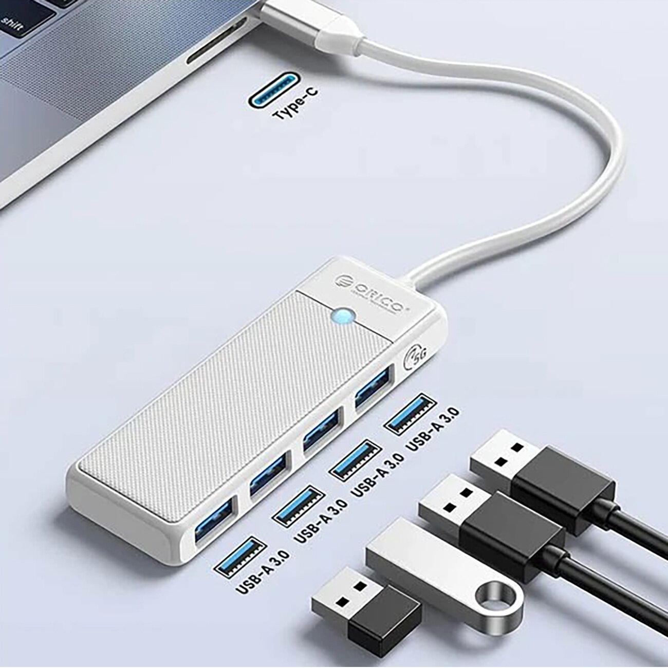 Na obrazie widoczny jest biały hub USB z czterema portami USB-A 3.0, podłączony do laptopa przez kabel USB-C. Na hubie znajduje się logo \'Orico\' oraz niebieska dioda, a obok leżą różne urządzenia USB.