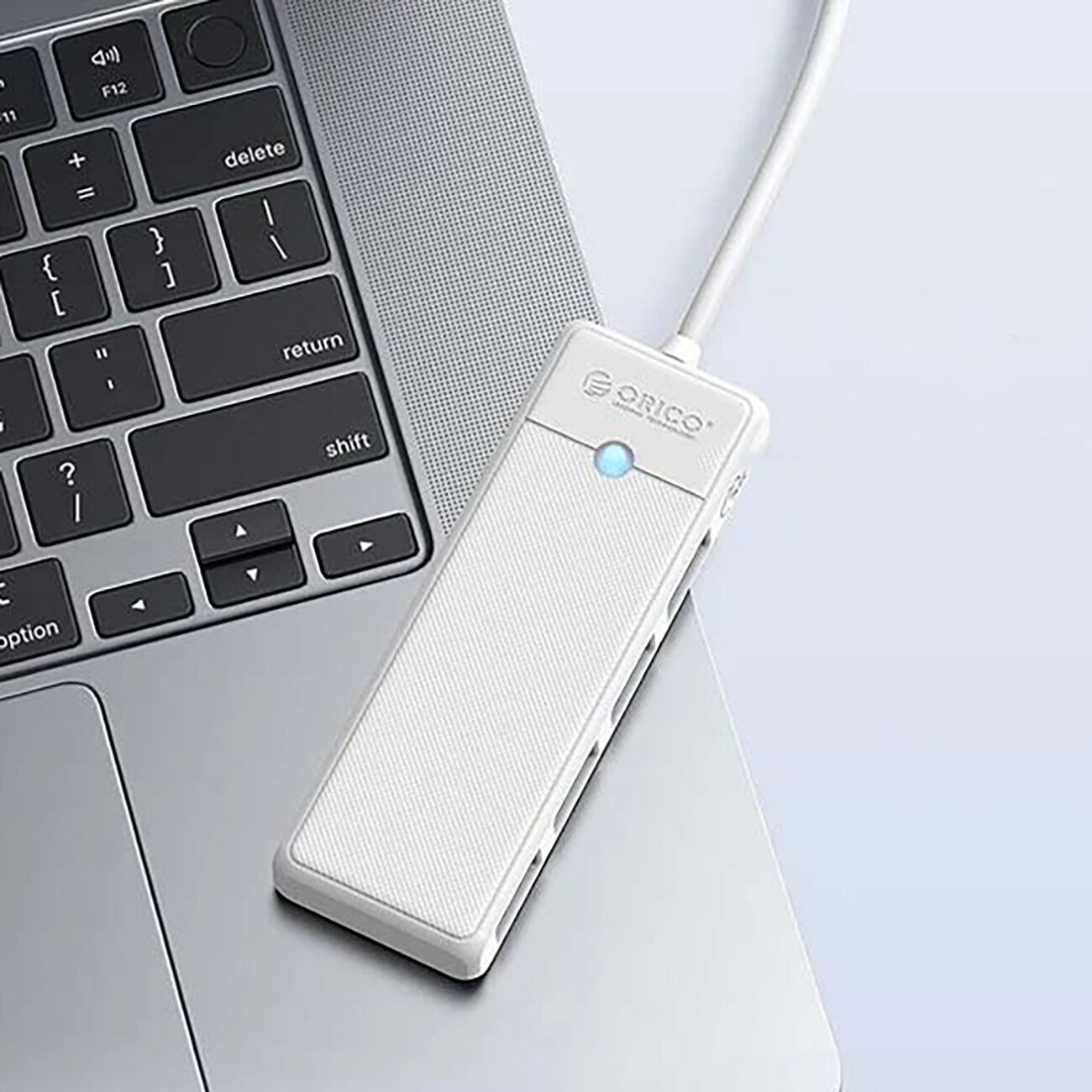Na obrazie widoczny jest biały hub USB z logo \'Orico\', podłączony do laptopa. Hub posiada cztery porty USB-A i świecący niebieski wskaźnik.