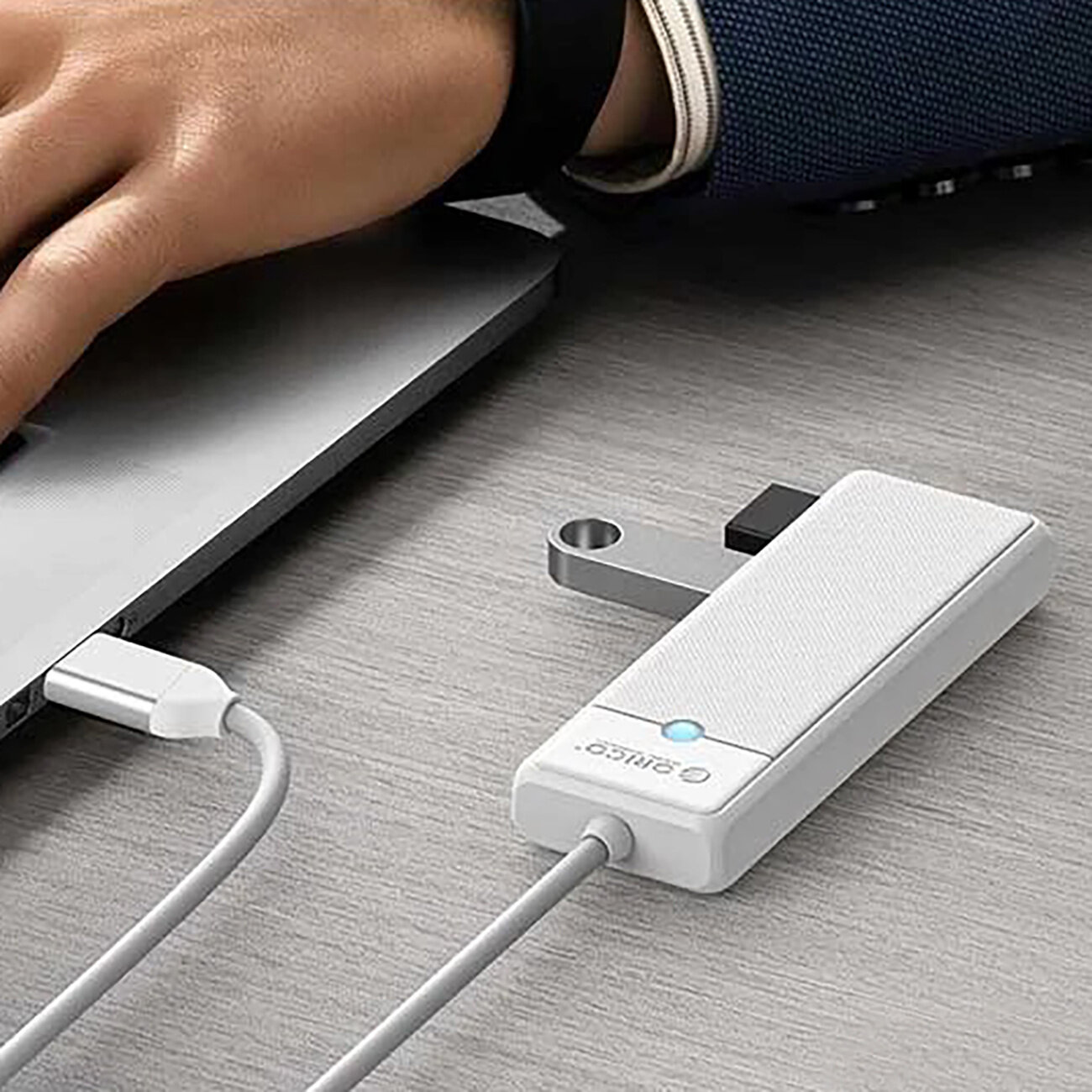 Na biurku znajduje się biały hub USB z logo \'ORICO\', podłączony do laptopa. Obok widoczna jest ręka osoby korzystającej z laptopa.