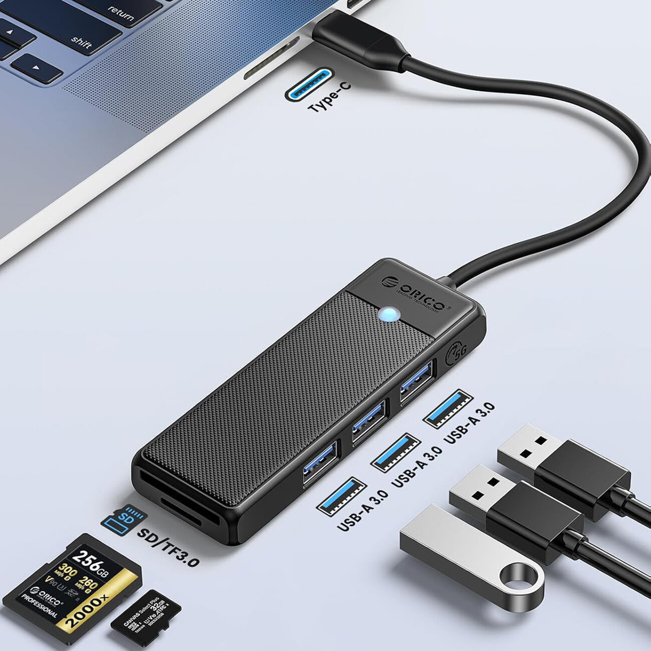 Na obrazie widoczny jest czarny hub USB z trzema portami USB-A 3.0 i czytnikiem kart microSD/SD, podłączony do laptopa przez kabel USB-C. Obok znajdują się karty pamięci i pendrive, a na hubie widnieje logo \'Orico\'.