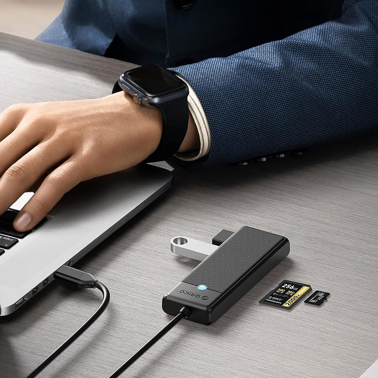 Na biurku leży czarny hub USB z logo \'Orico\', podłączony do laptopa. Obok znajdują się karty microSD i SD, a w tle widoczna jest ręka osoby w niebieskiej marynarce z zegarkiem na nadgarstku.