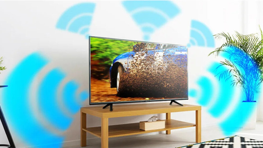 Telewizor stoi na drewnianym stoliku, a na ekranie widać samochód jadący po błotnistej drodze. W tle widoczne są niebieskie fale symbolizujące sygnał oraz rośliny doniczkowe.