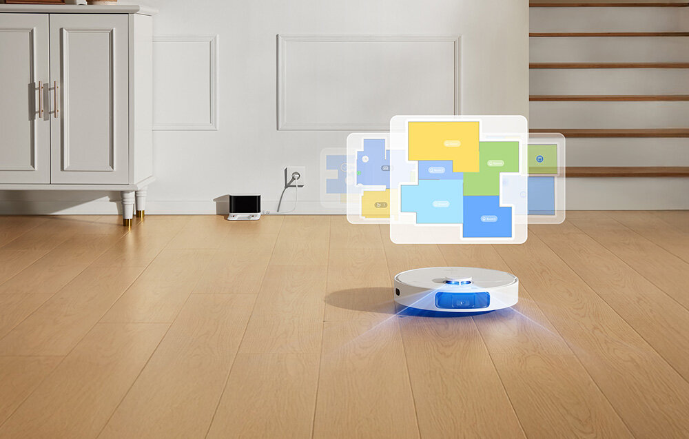 Na drewnianej podłodze znajduje się biały robot sprzątający z niebieskim podświetleniem. W tle widać szafkę, schody oraz stację dokującą podłączoną do gniazdka elektrycznego.
