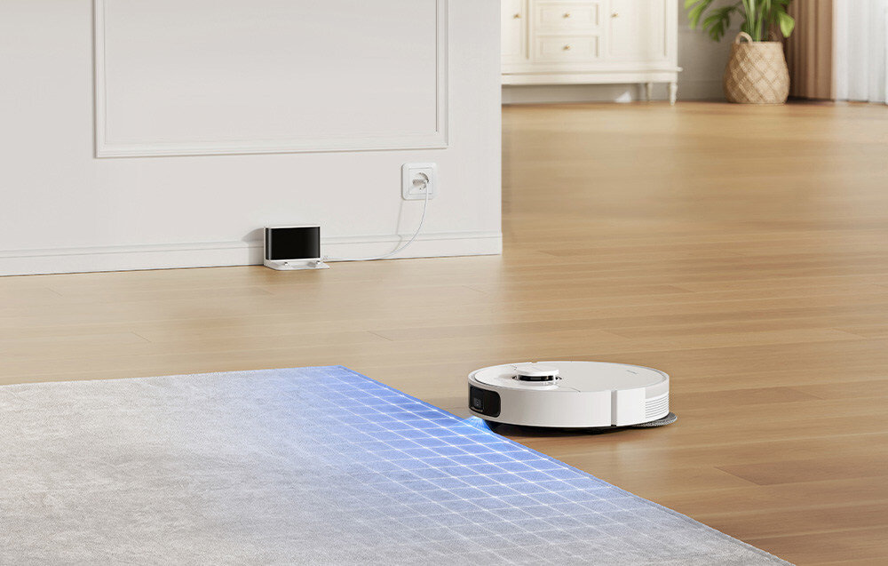 Na drewnianej podłodze znajduje się biały robot sprzątający, który zbliża się do dywanu z niebieską siatką świetlną. W tle widać stację dokującą podłączoną do gniazdka elektrycznego na ścianie.