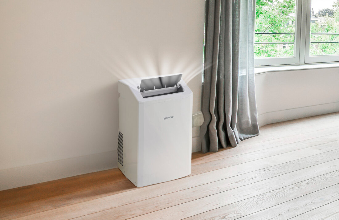 Biały klimatyzator przenośny z logo \'gorenje\' stoi na drewnianej podłodze przy ścianie. Obok znajduje się zasłona i okno z widokiem na zieleń.