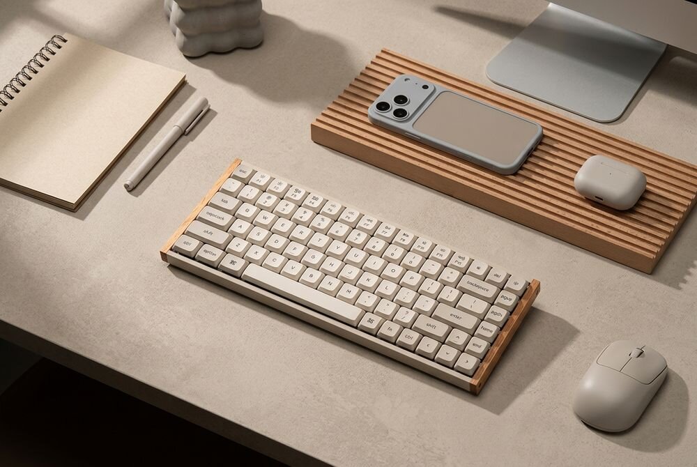 Na biurku znajduje się klawiatura z jasnymi klawiszami, obok której leży myszka, notatnik, długopis oraz smartfon w etui na drewnianej podstawce. W tle widoczny jest monitor i etui na słuchawki.