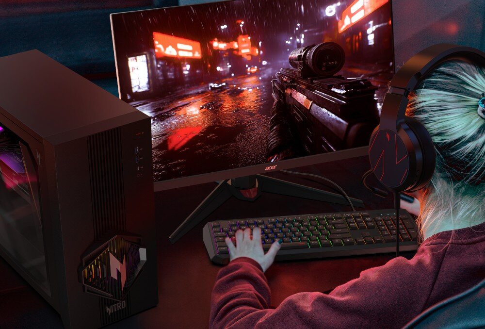 Osoba siedzi przed komputerem z dużym monitorem, na którym wyświetlana jest gra z motywem futurystycznego miasta. Obok monitora znajduje się obudowa komputera z widocznym logo \'Nitro\'.