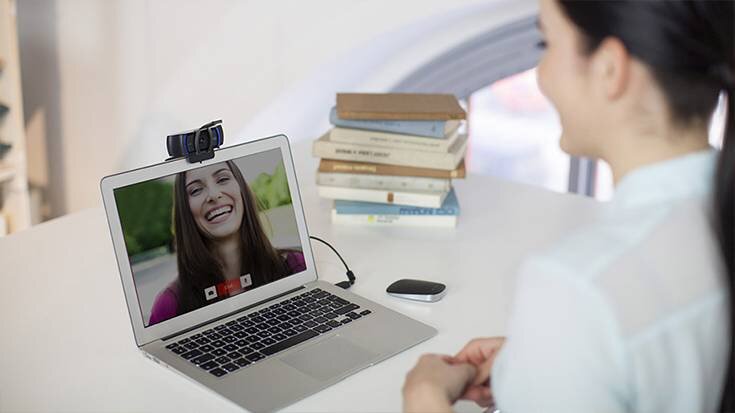 Na biurku stoi otwarty laptop z włączonym wideorozmową, na ekranie widać uśmiechniętą osobę. Na laptopie zamontowana jest kamera internetowa, a obok leży stos książek i małe urządzenie.
