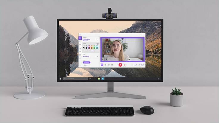 Na biurku znajduje się monitor z włączonym programem do wideokonferencji, na którym widać osobę. Na monitorze zamontowana jest kamera internetowa, obok stoi lampa, klawiatura, myszka i mała roślina w doniczce.