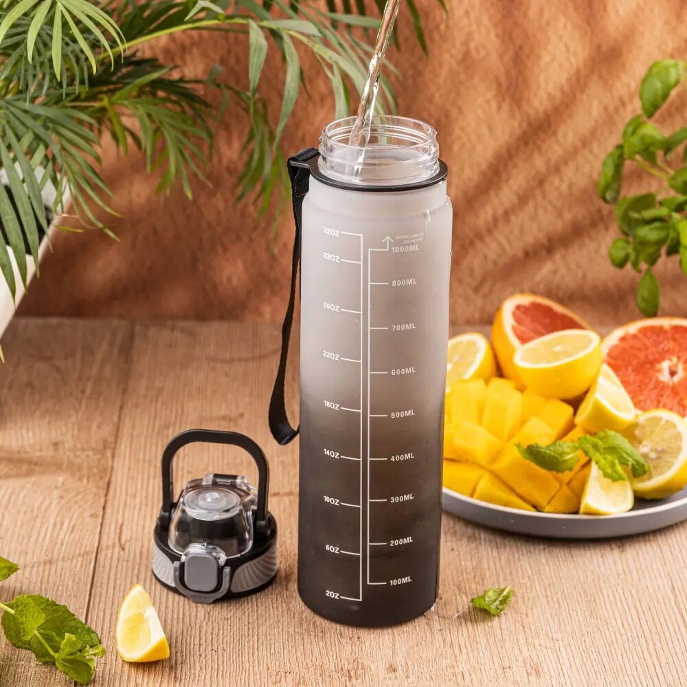 Na drewnianym stole stoi przezroczysty bidon z miarką od 100 ml do 1000 ml, obok leży zakrętka. W tle widoczne są plastry cytrusów i zielone liście.
