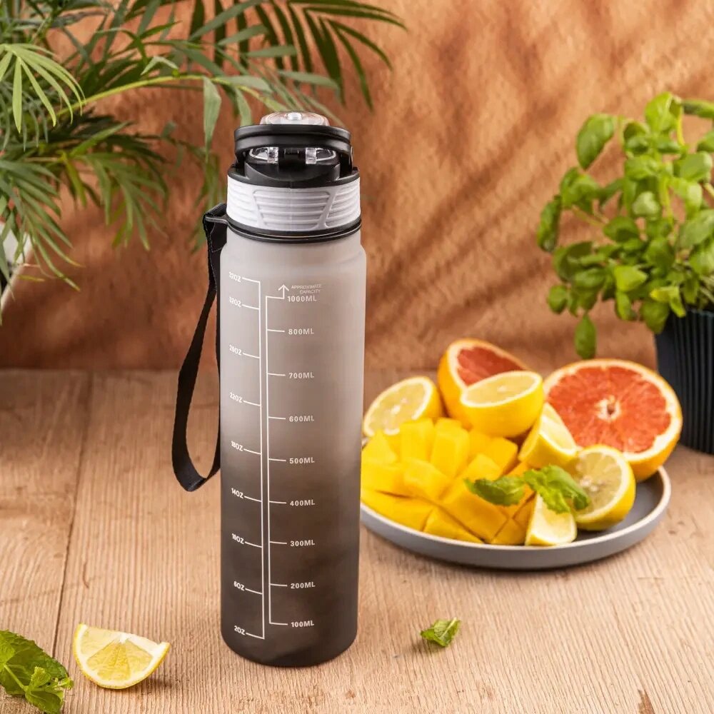 Na drewnianym stole stoi przezroczysty bidon z miarką do 1000 ml i czarną zakrętką. Obok znajduje się talerz z pokrojonymi owocami, takimi jak cytryny i grejpfruty, oraz zielona roślina w doniczce.