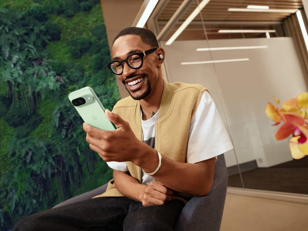 Osoba siedzi na krześle, trzymając w ręku zielony smartfon z logo \'G\'. W tle widoczna jest ściana z roślinnością oraz szklana przegroda.