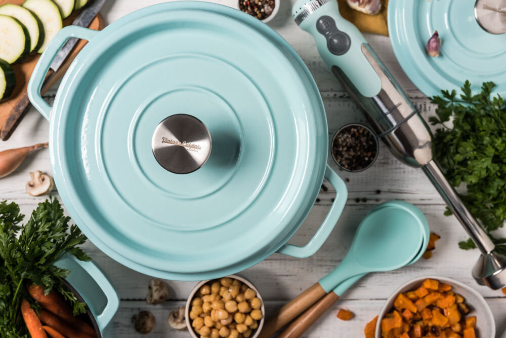 Na obrazie widoczny jest turkusowy garnek z pokrywką, na której znajduje się napis \'Vintage Cuisine\'. Wokół garnka rozmieszczone są różne składniki kulinarne, takie jak warzywa i przyprawy, oraz turkusowe akcesoria kuchenne.