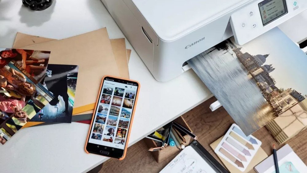 Na biurku znajduje się urządzenie wielofunkcyjne Canon, które drukuje kolorowe zdjęcie przedstawiające miasto nad wodą. Obok leży smartfon z wyświetlonymi miniaturami zdjęć oraz kilka wydrukowanych fotografii.