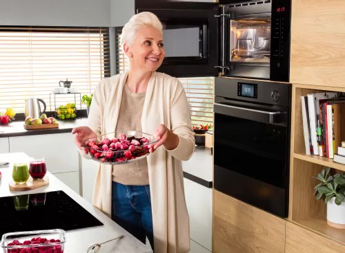 Kobieta trzymająca talerz z owocami stoi w nowoczesnej kuchni, obok otwartej kuchenki mikrofalowej w zabudowie. W tle widać różne owoce i sprzęty kuchenne na blacie.