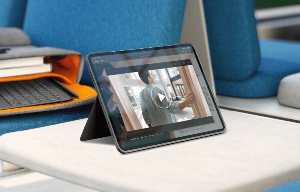 Tablet w etui stoi na stole, wyświetlając wideo z osobą w pomieszczeniu. Obok leży klawiatura i otwarta torba z książkami.