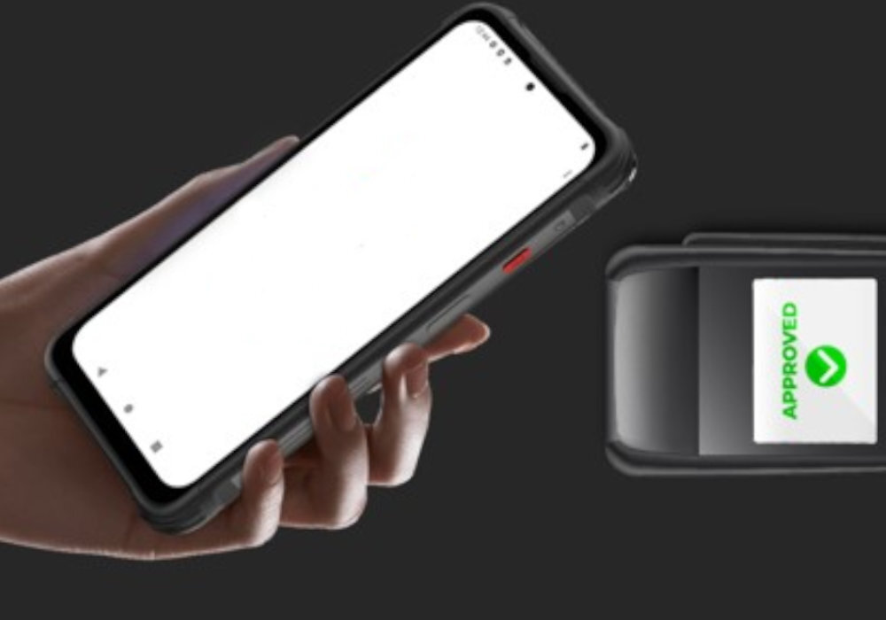 Osoba trzyma czarny smartfon z dużym ekranem, obok znajduje się urządzenie z wyświetlaczem pokazującym napis \'APPROVED\' z zielonym symbolem. Tło jest ciemne, a smartfon ma widoczny czerwony przycisk na boku.