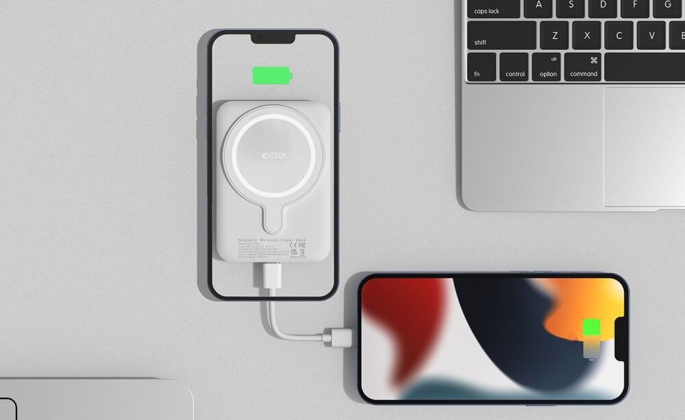 Na obrazie widać biały powerbank z logo TECH-PROTECT, podłączony do smartfona, który pokazuje ikonę ładowania. Obok znajduje się laptop oraz drugi smartfon z kolorowym ekranem i ikoną ładowania.