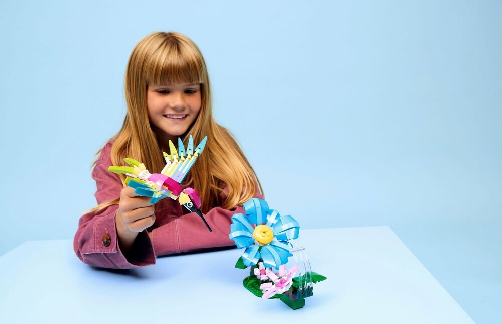 Dziewczynka trzyma kolorowy model kolibra z klocków LEGO, obok znajduje się niebieski kwiat również z klocków LEGO, na jasnoniebieskim tle.