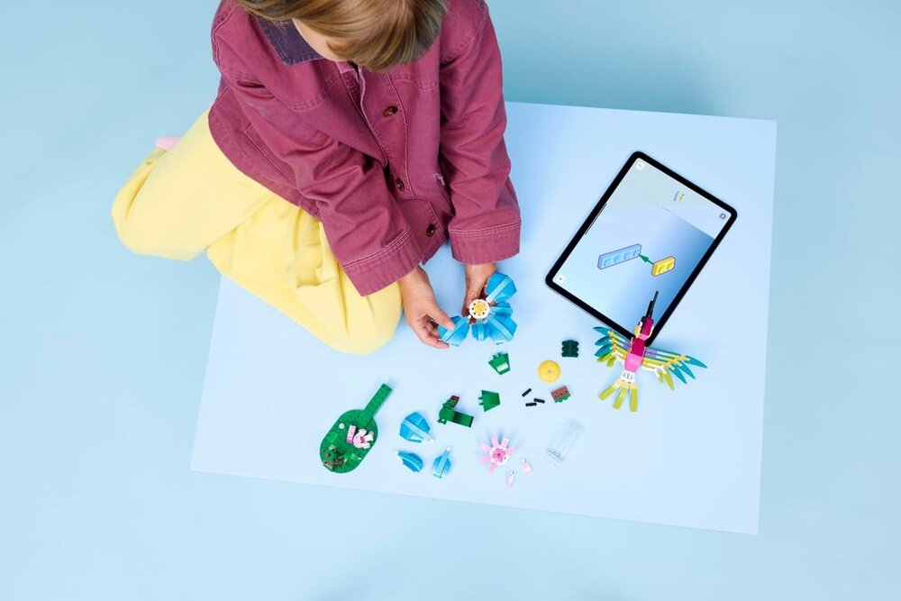 Dziecko siedzi na podłodze i bawi się zestawem klocków LEGO, obok leży tablet z instrukcją. Na białej powierzchni rozłożone są różnokolorowe elementy LEGO, w tym złożony kolorowy ptak.