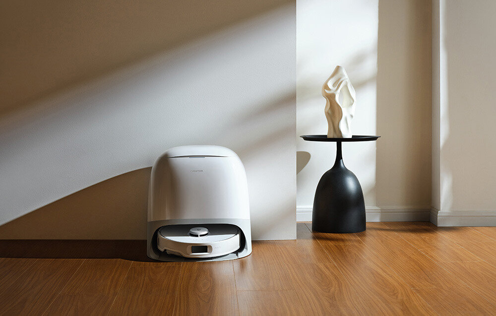 Na drewnianej podłodze stoi robot sprzątający w stacji dokującej obok czarnego stolika z białą rzeźbą. Ściana w tle jest jasna, a na nią padają cienie.