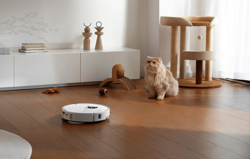 Na drewnianej podłodze znajduje się biały robot sprzątający. Obok stoi kot, a w tle widać meble i zabawki dla kota.
