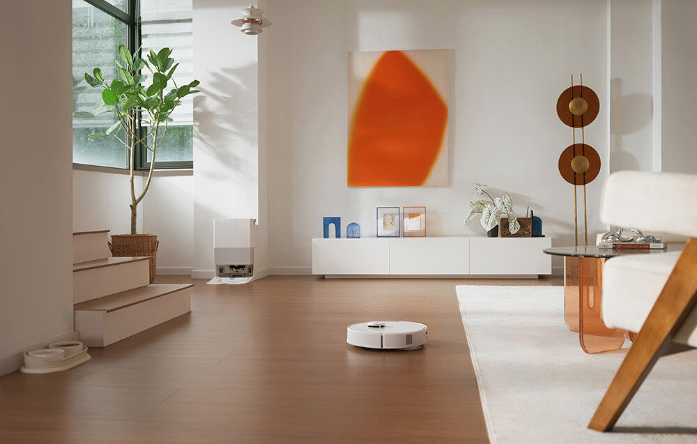 W nowoczesnym salonie z drewnianą podłogą znajduje się biały robot sprzątający. W tle widać białą szafkę z dekoracjami i dużą abstrakcyjną grafikę w pomarańczowych odcieniach na ścianie.