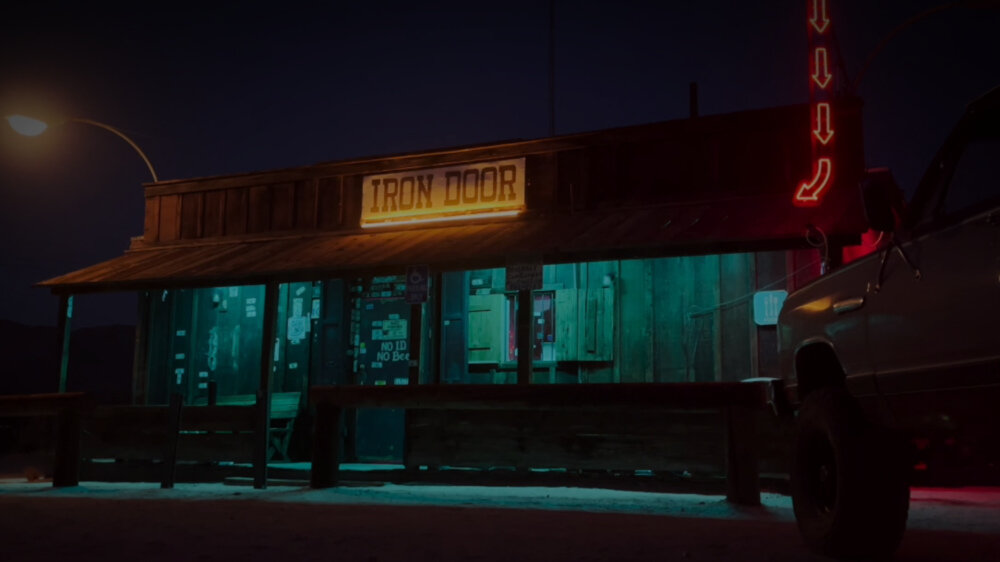 Nocna scena przedstawia drewniany budynek z neonowym napisem \'IRON DOOR\'. Obok budynku stoi samochód, a w tle widać latarnię uliczną.
