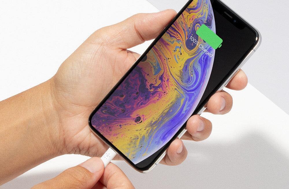 Osoba trzyma telefon z kolorowym wyświetlaczem, na którym widoczny jest napis \'100% Charged\'. Do telefonu podłączony jest biały kabel USB-C.