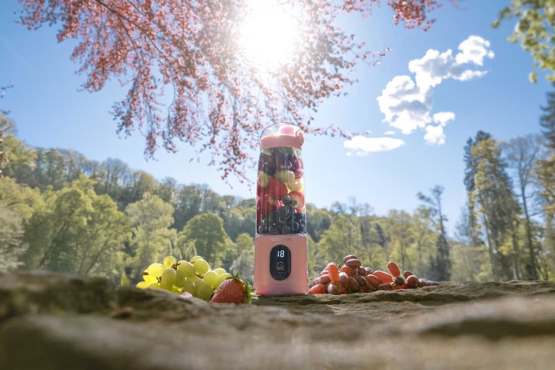 Na kamiennej powierzchni stoi różowy blender wypełniony owocami, obok leżą winogrona i truskawki. W tle widać drzewa i niebo z chmurami, a słońce świeci jasno.