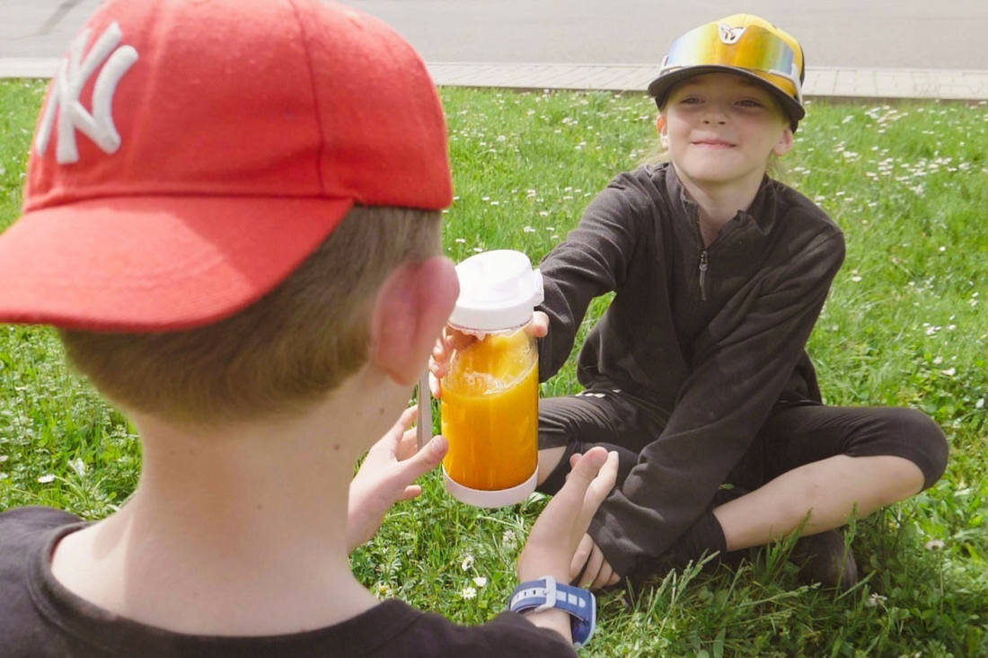 Dwoje dzieci siedzi na trawie, jedno z nich trzyma w ręku blender z pomarańczowym napojem. Dziecko z przodu ma na głowie czerwoną czapkę z białym logo \'NY\'.