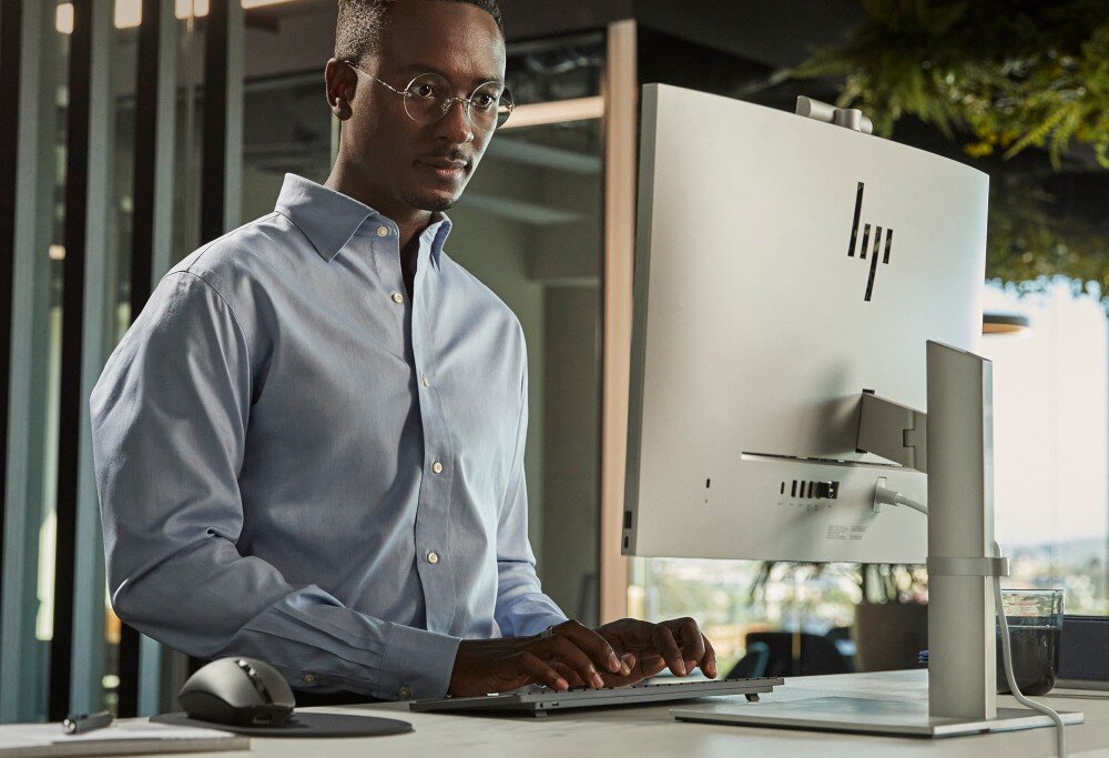 Osoba stoi przy biurku, używając komputera z logo HP na monitorze. W tle widoczne są elementy biurowe i roślinność.