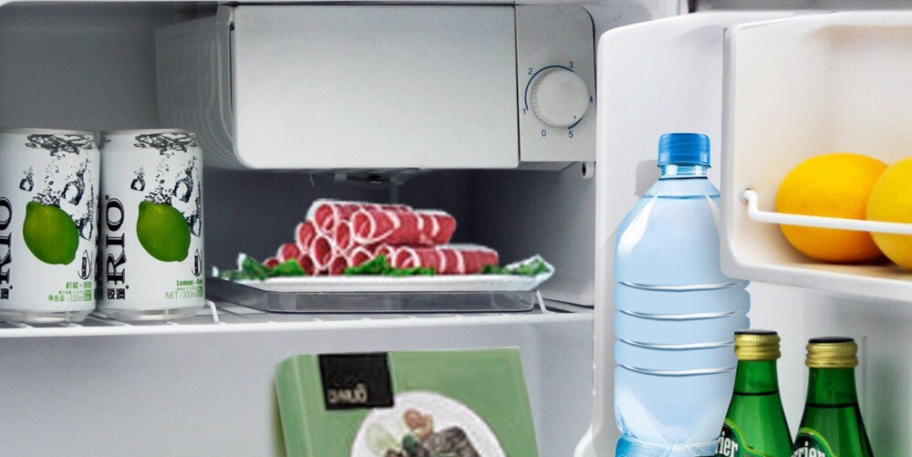 Wnętrze lodówki z widocznymi produktami: dwie puszki napoju z napisem \'RIO\', talerz z plastrami mięsa, butelka wody, dwie butelki Perrier oraz cytryny na drzwiach.