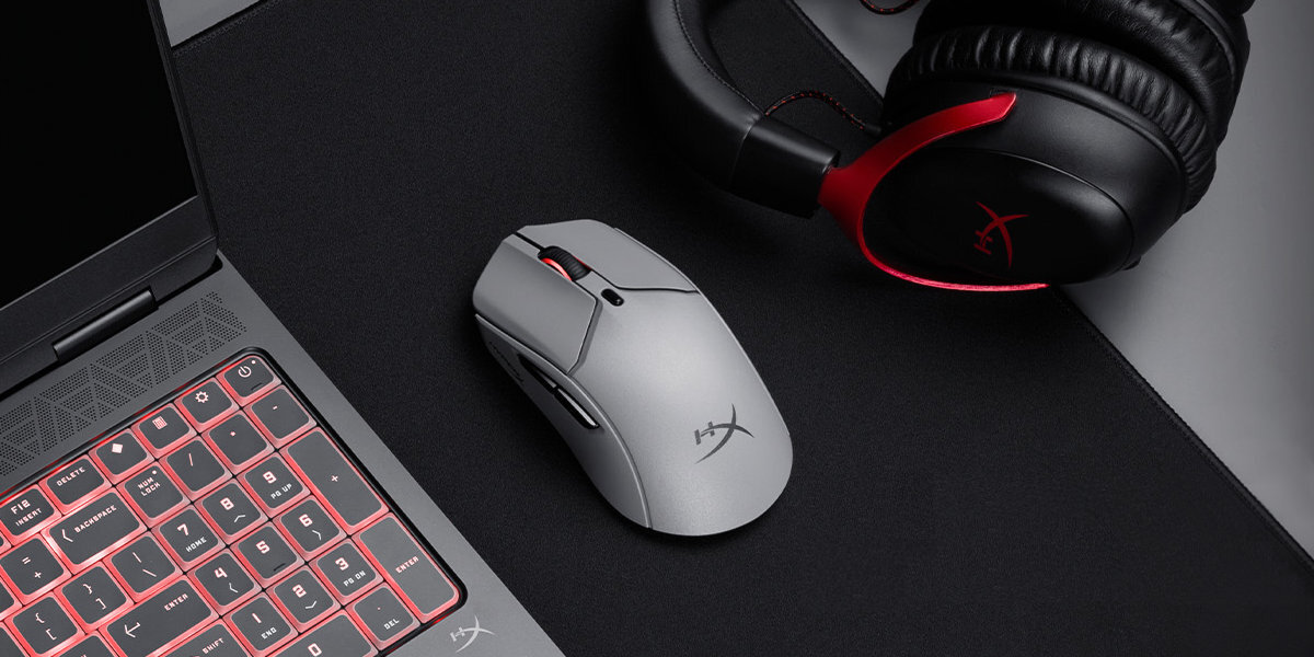 Na czarnym podkładzie znajduje się szara mysz komputerowa z logo \'HyperX\', obok której leżą słuchawki z czerwonymi akcentami oraz laptop z podświetlaną klawiaturą.