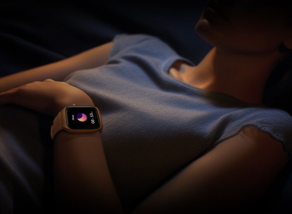 Osoba leży w ciemnym otoczeniu, nosząc smartwatch z wyświetlaczem pokazującym ikonę księżyca i czas 08:55. Smartwatch ma jasny pasek i kwadratowy ekran.