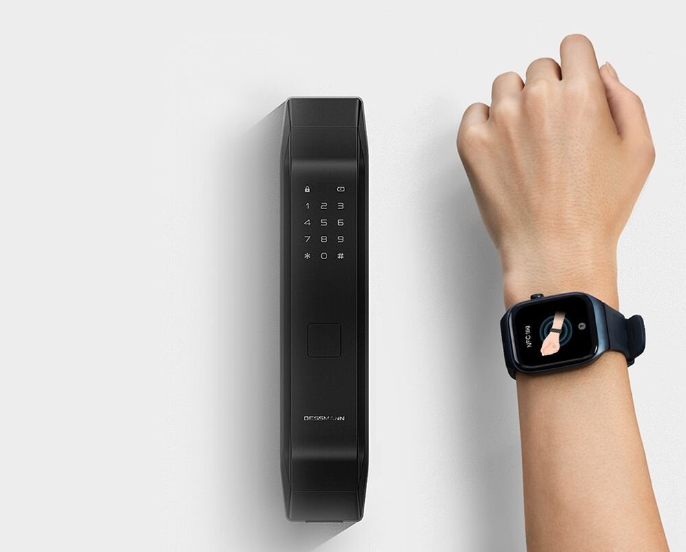 Na obrazie widoczny jest czarny smartwatch na nadgarstku osoby oraz czarny panel z klawiaturą numeryczną i symbolem kłódki. Na ekranie zegarka wyświetla się obraz dłoni.